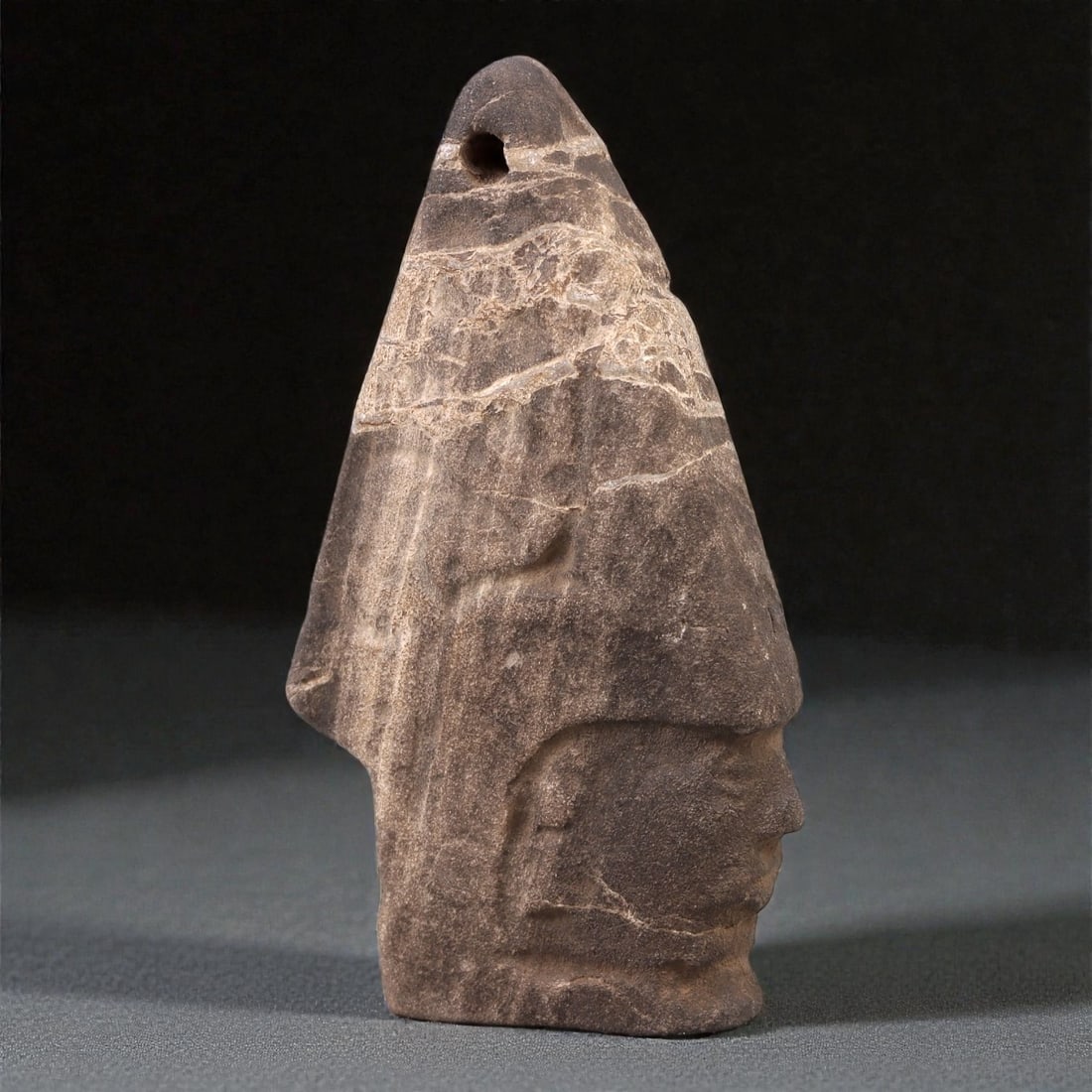 A Mesoamerican Stone Head Pendant, Pre-Columbian Period, 38R: Carved from a single piece of fine-grained dark brown stone with visible natural veining and surface fissures, the object represents a highly stylized human head rendered in profile. The form is marke