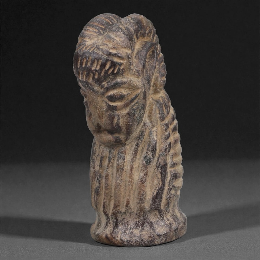 A Syro-Hittite Schist Ram-Head Idol, Early Iron Age, 11th-9th Century BCE, 90LJ: Carved from mottled grey-brown schist, the object is fashioned as a stylized ram's head set upon a cylindrical base. The facial features are rendered with deeply recessed eyes, a short muzzle, and a s