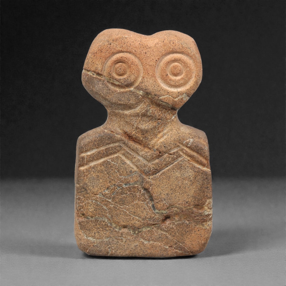 A Northern Mesopotamian Stone Eye Idol, 4th Millennium BCE, 46AC: Carved from a warm brown stone with lighter veining, the figure is fashioned with a rectangular lower body and a bifurcated upper section bearing two concentric recessed circular eyes. The neck area i