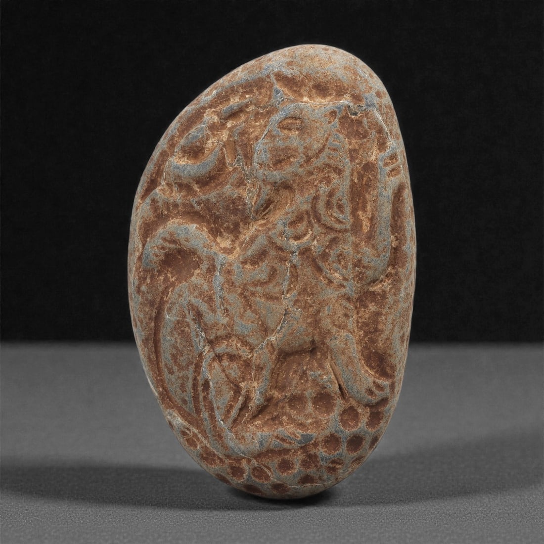 A Byzantine Stone Relief Plaque with Lion Figure, 11th-12th Century, 24B: Carved from a reddish-brown stone with pale blue-grey surface accretions, the plaque is shaped into a vertically elongated oval. The relief depicts a lion in profile, shown rearing with its forelegs r