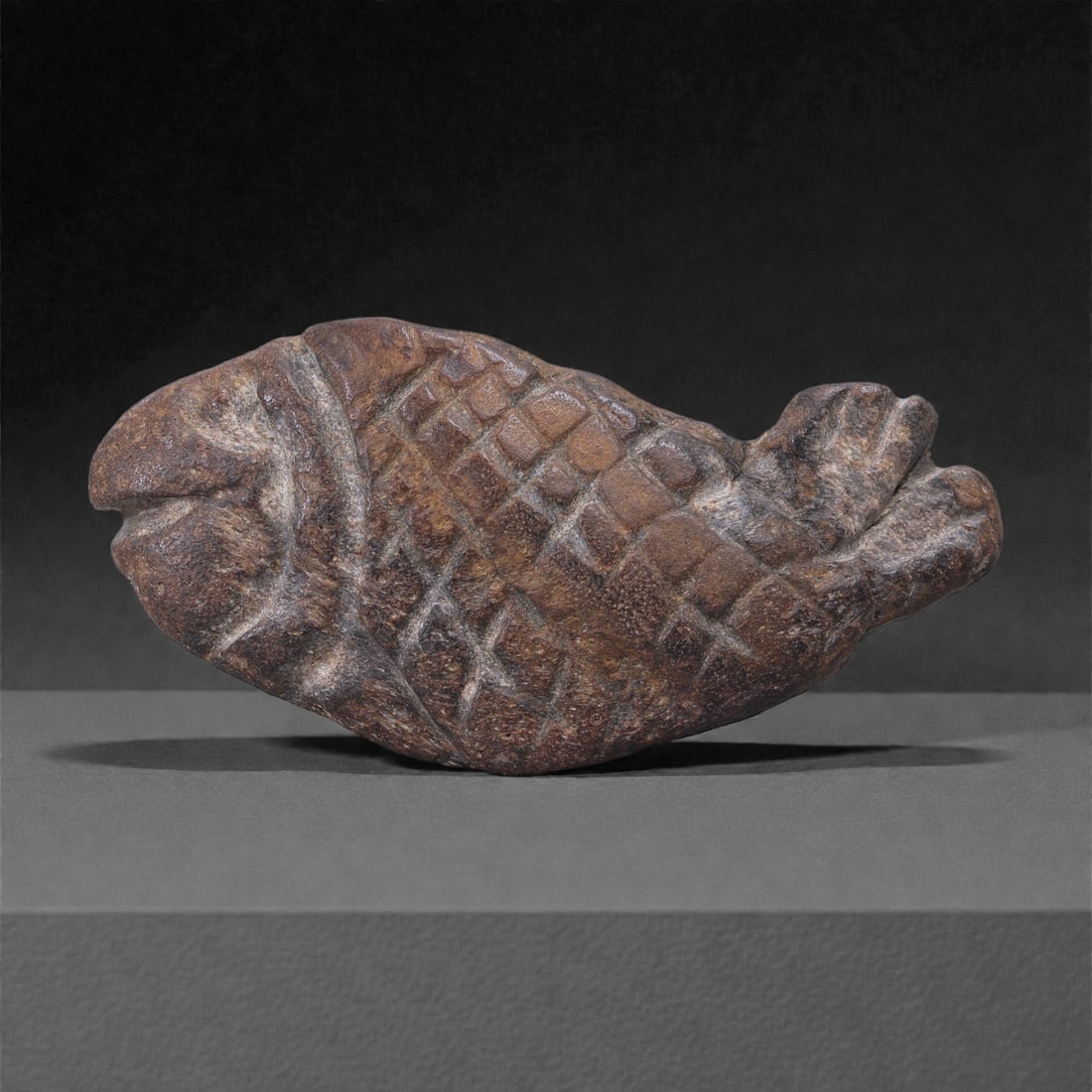 A Near Eastern Stone Fish Effigy, 3rd-2nd Millennium BCE, 99M: Carved from a dense brown stone with lighter mineral veining, the object represents a fish shown in profile with a broad, flattened body. The head is schematically formed with a projecting snout and s