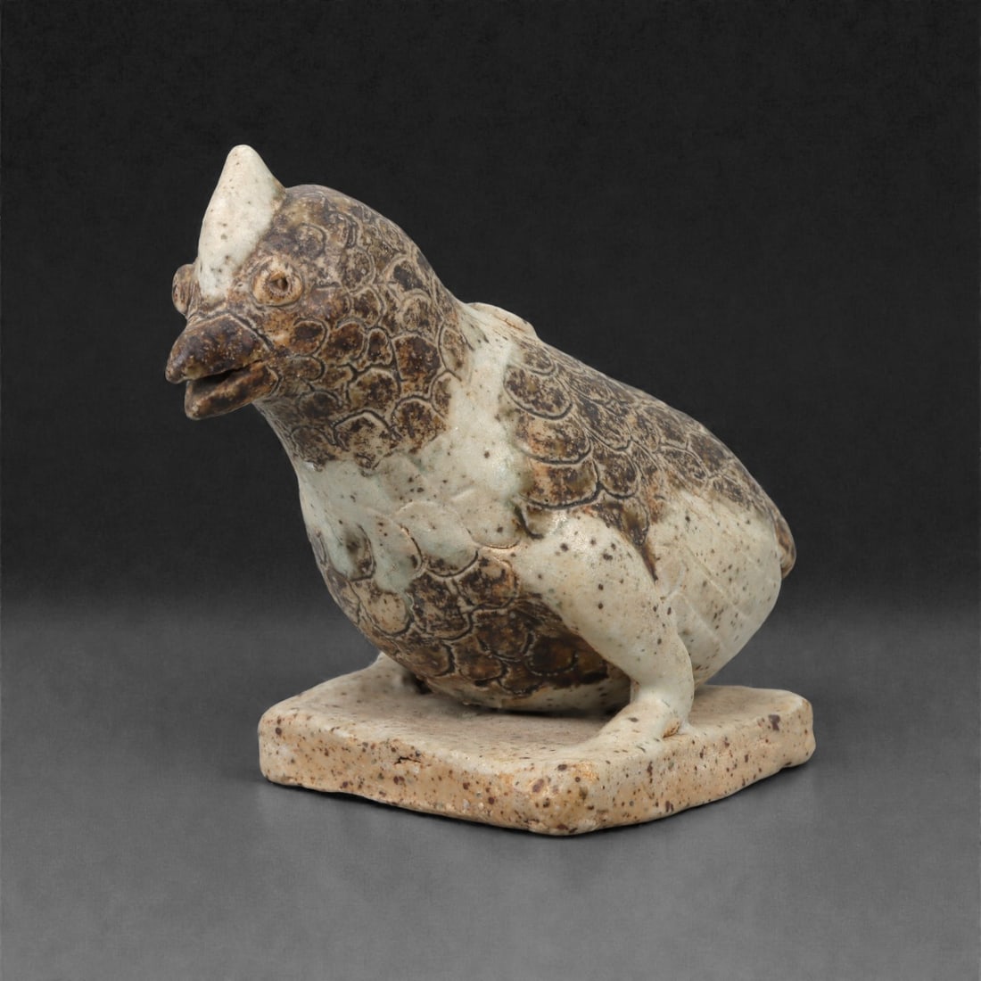 An Islamic Ceramic Bird Effigy, Seljuk Period, 12th-13th Century CE, 19LN: Modelled in a buff ceramic fabric with a mottled glazed surface, the figure represents a bird seated on a flat square base. The body is rounded and covered with scale-like incised motifs, while the he