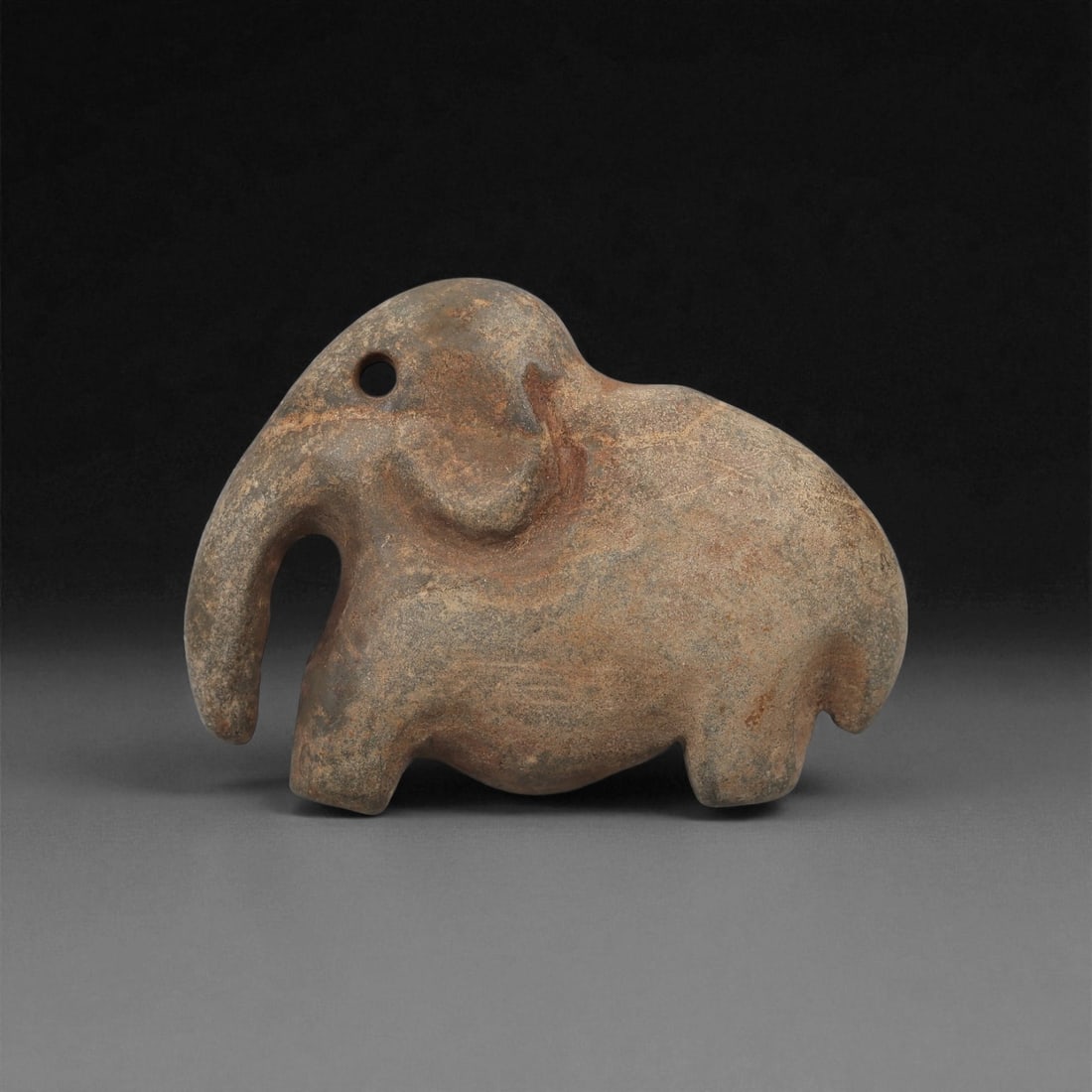 A Near Eastern Stone Elephant Figurine, 3rd-2nd Millennium BCE, 12LA: Carved from grey-brown stone with lighter surface mottling, the figure depicts an elephant in profile with a rounded body supported on four short legs. The head is modeled with a drilled circular eye,