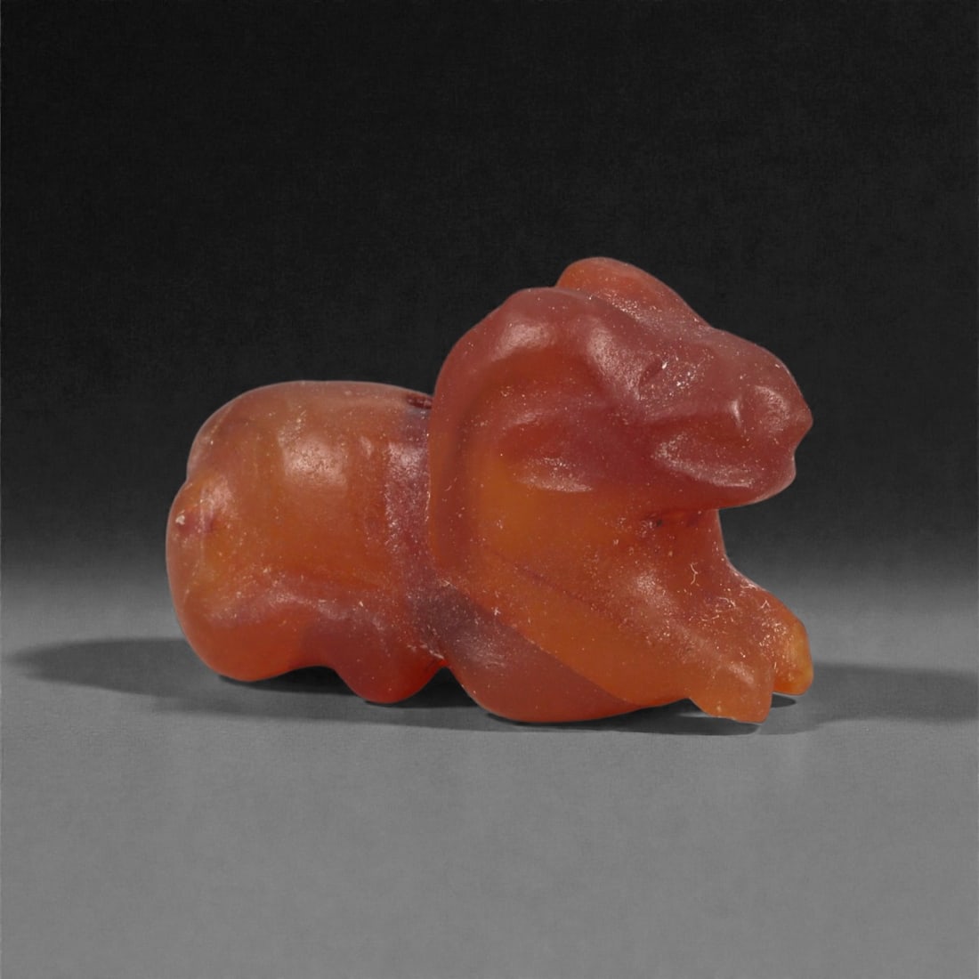 A Near Eastern Carnelian Lion Amulet, 1st Millennium BCE, 80LG: Carved from translucent orange carnelian with natural internal banding, the figure is modeled in the round as a recumbent animal with a compact body and a slightly raised head. The facial features are