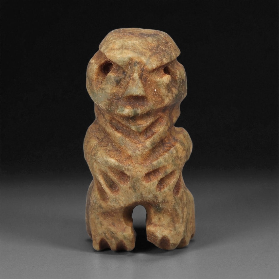 A Taino Carved Stone Idol, Late Ceramic Age, circa 1200-1500 CE, 94LE: Carved from mottled brown stone with greenish surface accretion, the figure is modeled standing with short legs separated at the base. The head is rounded with recessed circular eyes, a triangular nos