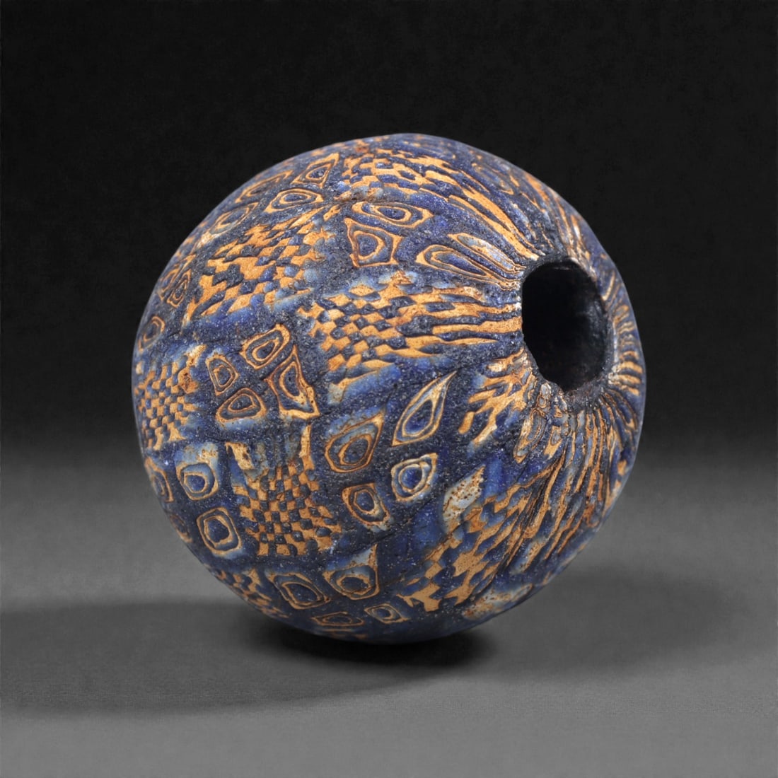 Ancient Egypto-Roman Mosaic Glass Bead - Feathered Eye-Cane Sphere, 42J: Worked in multicolored mosaic glass, this spherical bead displays an elaborate combination of elongated eye motifs and finely tessellated checkerboard canes arranged in radiating registers across a de