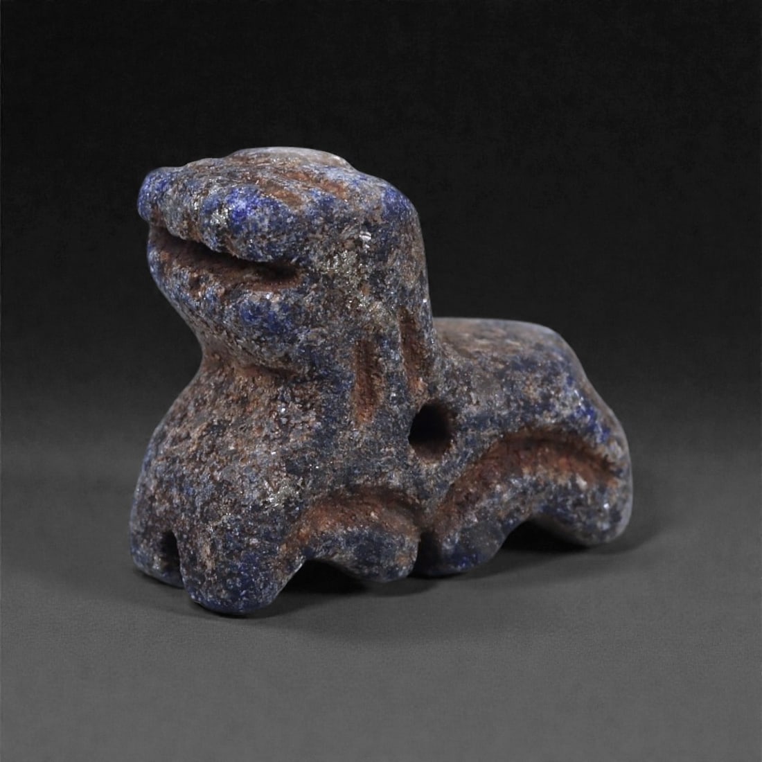 A Bactrian Lapis Lazuli Recumbent Animal Amulet, Late 3rd-Early 2nd Millennium BCE, 4N: Carved from lapis lazuli with mottled blue coloration and brownish mineral inclusions, the amulet is modeled as a recumbent quadruped with the head raised and the forelegs extended forward. The head i