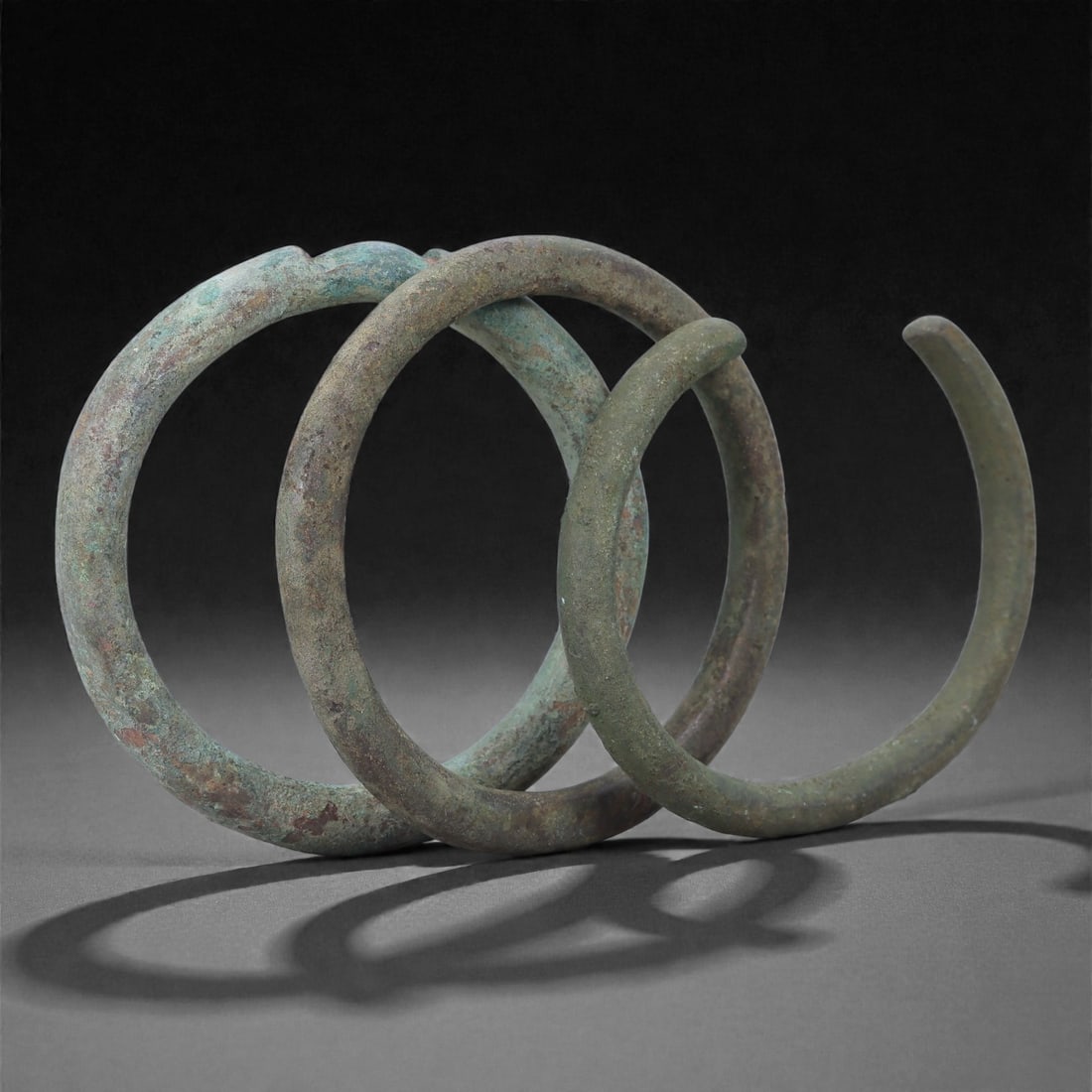Roman Bronze Bracelets, 1st-3rd Century CE, 25H: Cast in bronze with rounded circular profiles, the group comprises three open bracelets exhibiting green and brown patination with localized surface accretions consistent with burial conditions. Each 