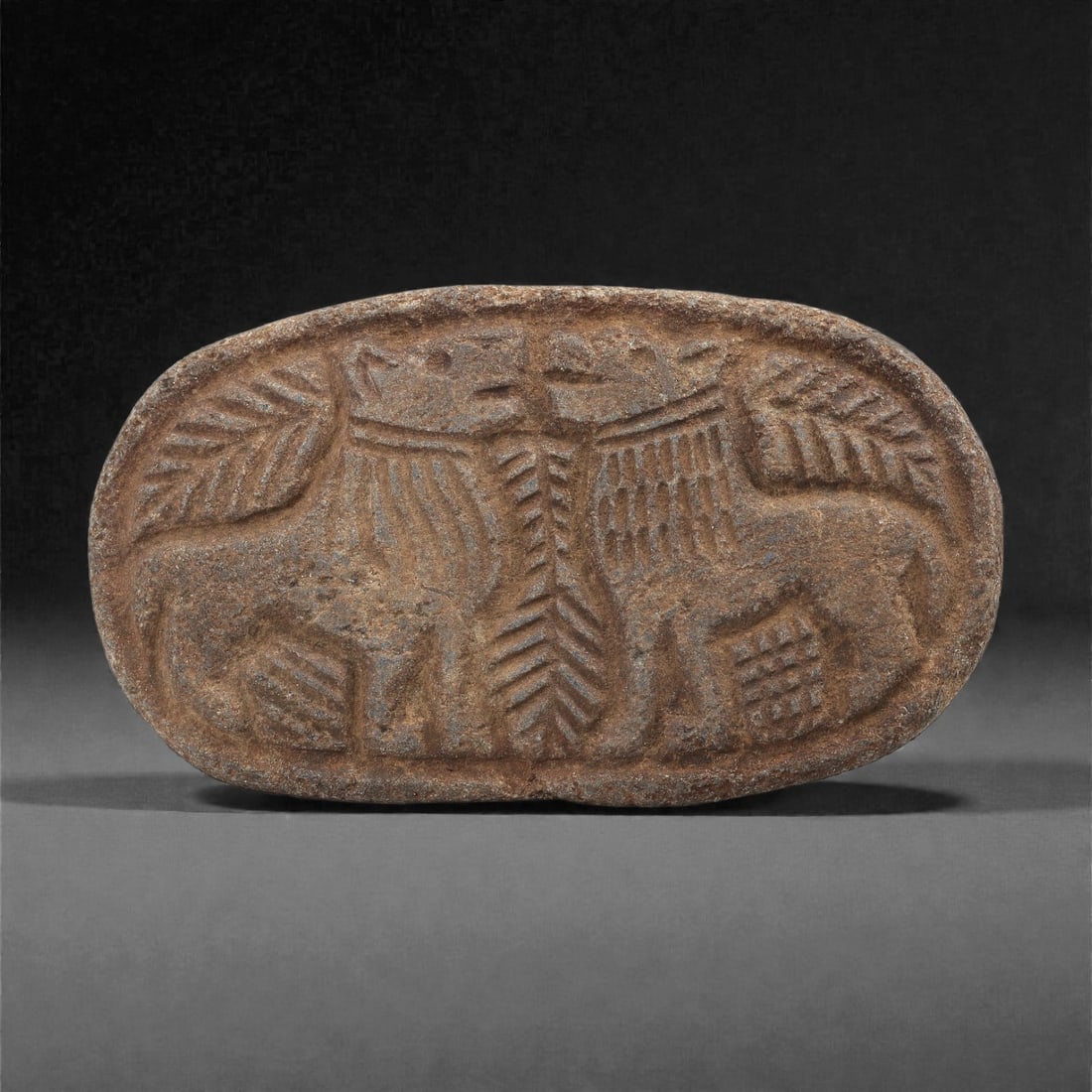 A Sasanian Lapis Lazuli Oval Plaque with Confronted Animals, 3rd-7th Century CE, 84LQ: Carved from lapis lazuli with a brown archaeological surface accretion, the oval plaque is worked in shallow relief with a pair of confronted quadrupeds flanking a central vertical motif formed by par