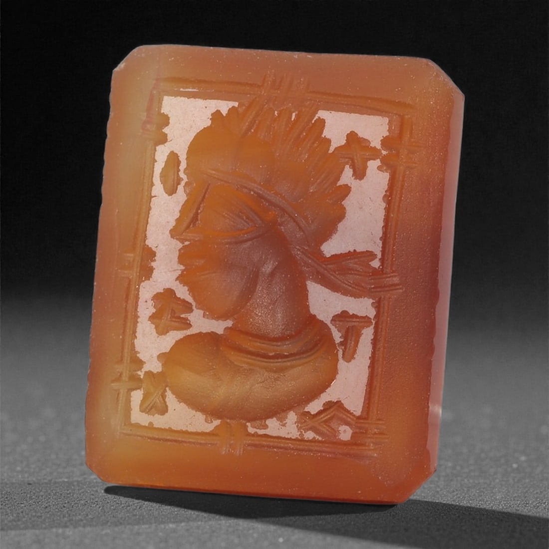 A Sasanian Carnelian Intaglio, 3rd-7th Century CE, 60LS: Carved from orange-brown carnelian, the gem is shaped into a rectangular plaque with faceted edges and engraved in intaglio with a male bust in left profile. The figure wears a radiating headdress wit