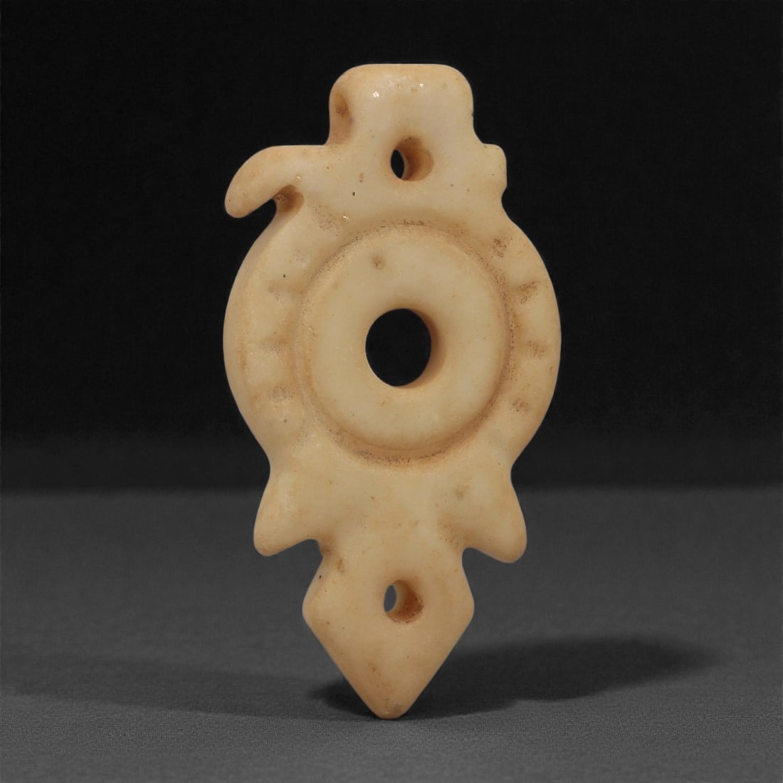 A Sino-Mongolian Jade Amulet, Medieval Period, 10th-14th Century CE, 17LQ: Carved from pale cream-colored jade with a smooth matte surface, the amulet is shaped around a centrally pierced circular body framed by a raised ring. The outer contour includes three short projectio