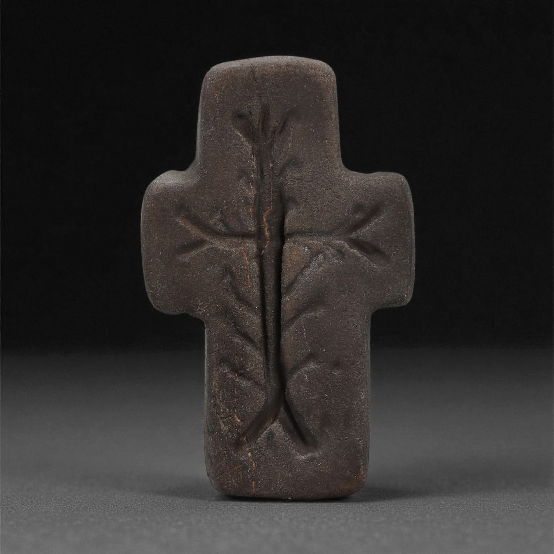 A Byzantine Stone Cross, 6th-8th Century CE, 9LD: Carved from a dark brown stone with a fine, matte surface, the object is shaped as a cross with rounded terminals. The front bears a shallow incised design featuring a vertical shaft intersected by a 