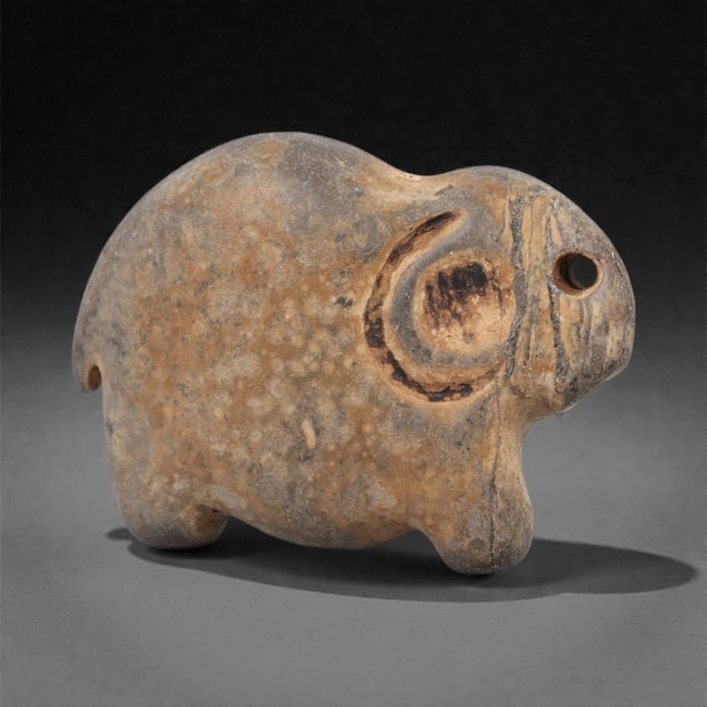 An Egyptian Stone Elephant Pendant, Predynastic Period, 4th Millennium BCE, 67LN: Carved from mottled brown stone with lighter surface accretions, the pendant represents a compact elephant with a rounded body, short projecting legs, and a downward-curving trunk. The eye is rendered