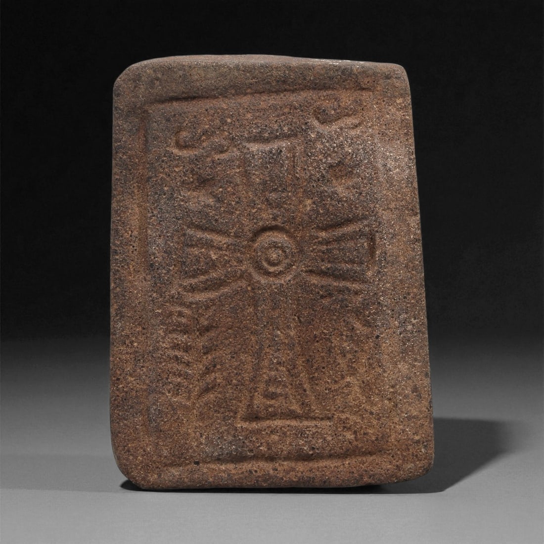 An Early Christian Stone Carved Cross Plaque, 5th-7th Century, 59LD: Carved from a coarse brown stone with a granular surface, the object is shaped as a rectangular plaque with a slightly rounded upper edge. The front face bears an incised cross with flared arms framed