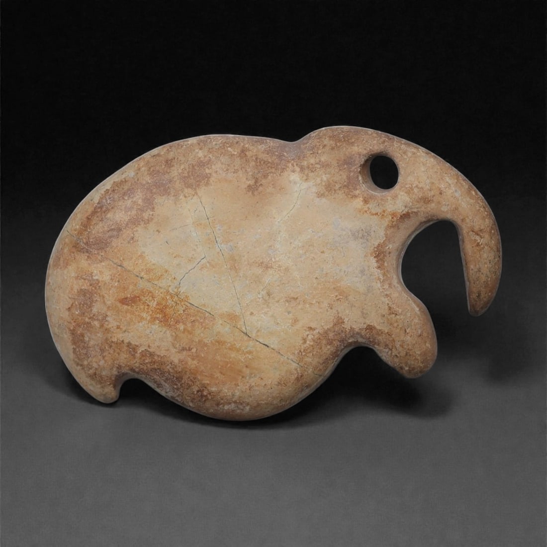 A Near Eastern Stone Elephant Pendant, 3rd-2nd Millennium BCE, 91LB: Carved from a pale stone with surface inclusions, the object is shaped in profile to depict an elephant with a rounded body and a pronounced curving trunk. A circular perforation is placed near the to