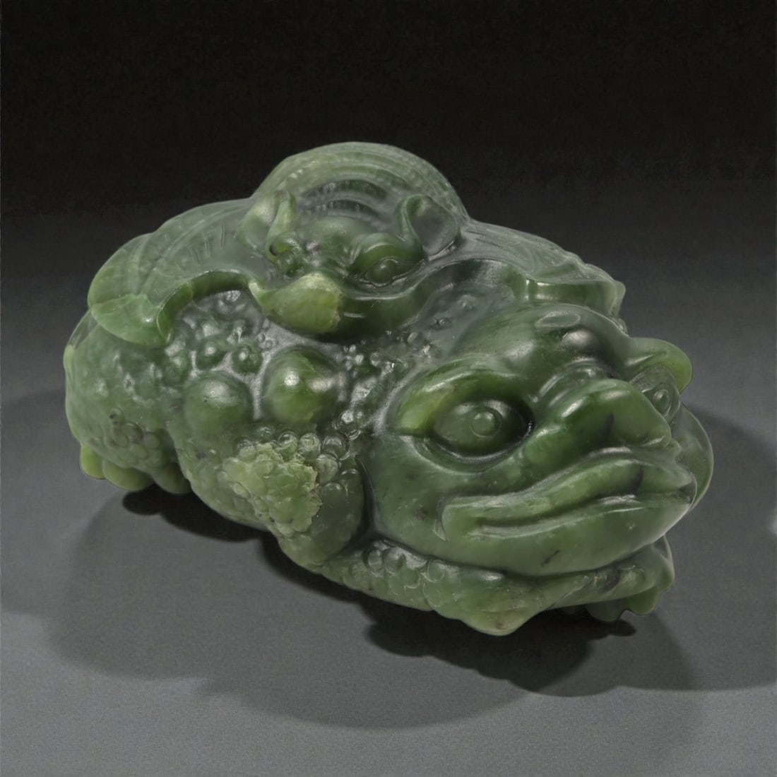 A Chinese Nephrite Jade Money Toad Figure, 20th Century, 93FB: Carved from dark green nephrite jade with areas of lighter mottling and natural inclusions, the figure represents a three-legged toad shown recumbent with the head turned slightly to one side. The bod