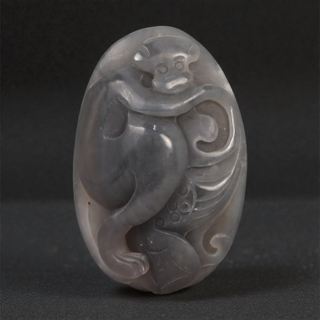 A Chinese Jade Monkey Plaque, 20th Century, 57YA: Carved from translucent gray jade with subtle tonal variation and areas of internal clouding, the object is formed as an oval plaque carved in low to moderate relief. The composition depicts a monkey