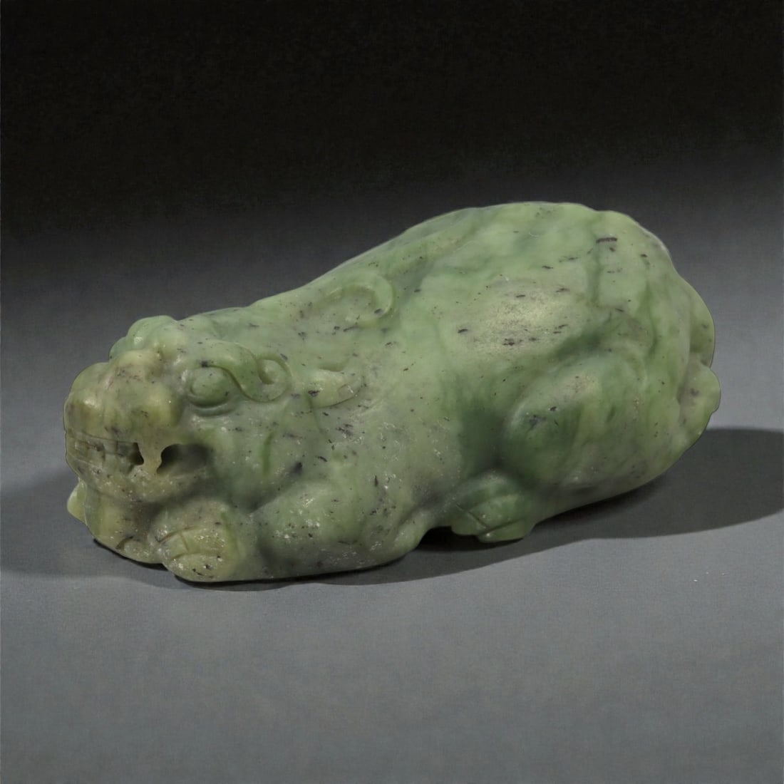 A Chinese Nephrite Jade Recumbent Mythical Beast, 20th Century, 95FB: Carved from mottled green nephrite jade with darker speckled inclusions, the figure represents a recumbent mythical beast shown resting with its body elongated and limbs tucked beneath. The head is ca