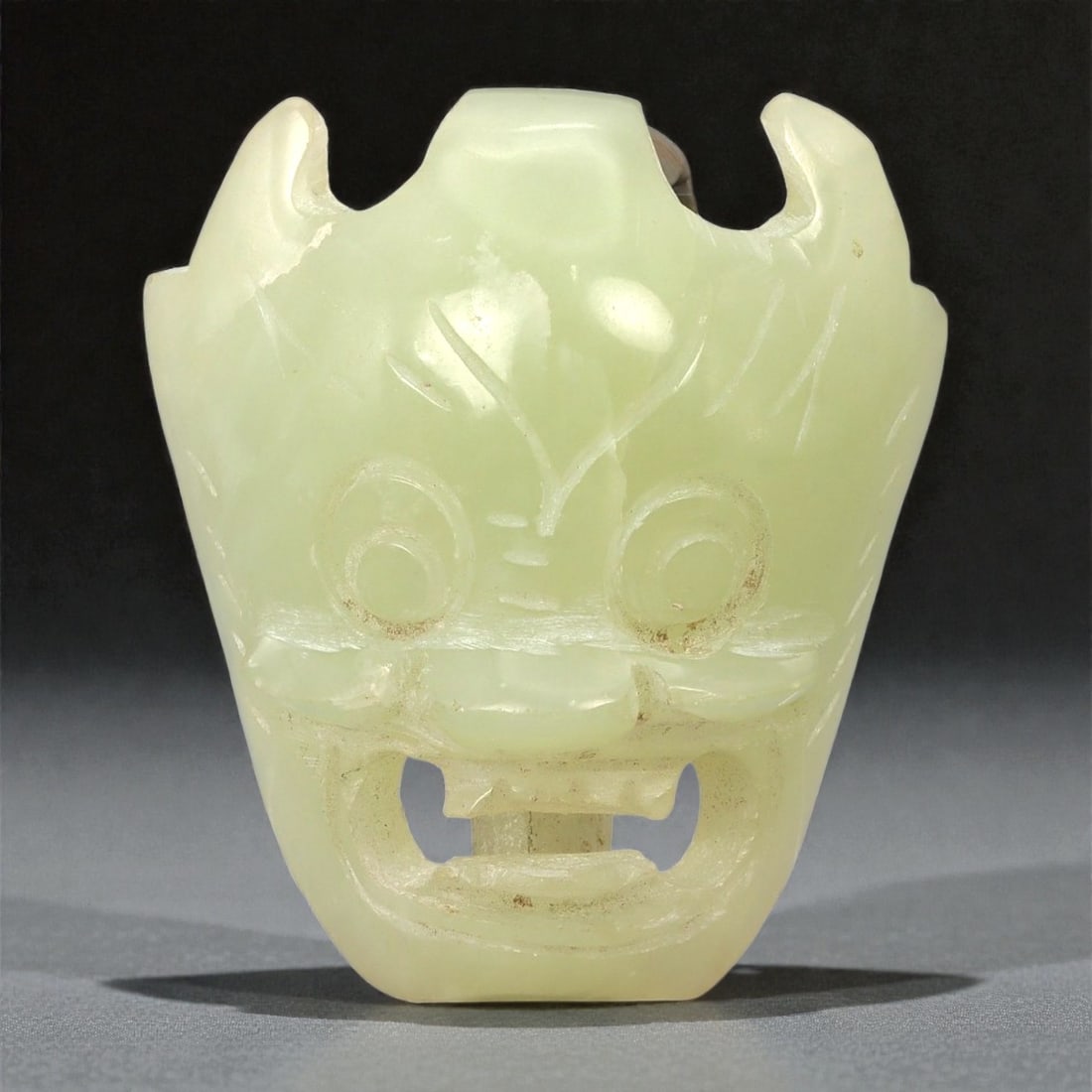 A Chinese Hetian Nephrite Jade Mask-Form Belt Buckle, 20th Century, 28YB: Carved from pale celadon Hetian nephrite with even translucency and a soft, waxy polish, the buckle is worked in the form of a stylized mask head. The face is symmetrically rendered with large circula