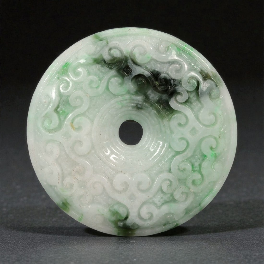 A Chinese Jadeite Jade Bi Disk, 19th-20th Century, 10FD: Carved from translucent jadeite jade of pale celadon tone with areas of brighter green and darker mineral concentrations, the disk is worked in a flattened circular form with a central perforation. Bo