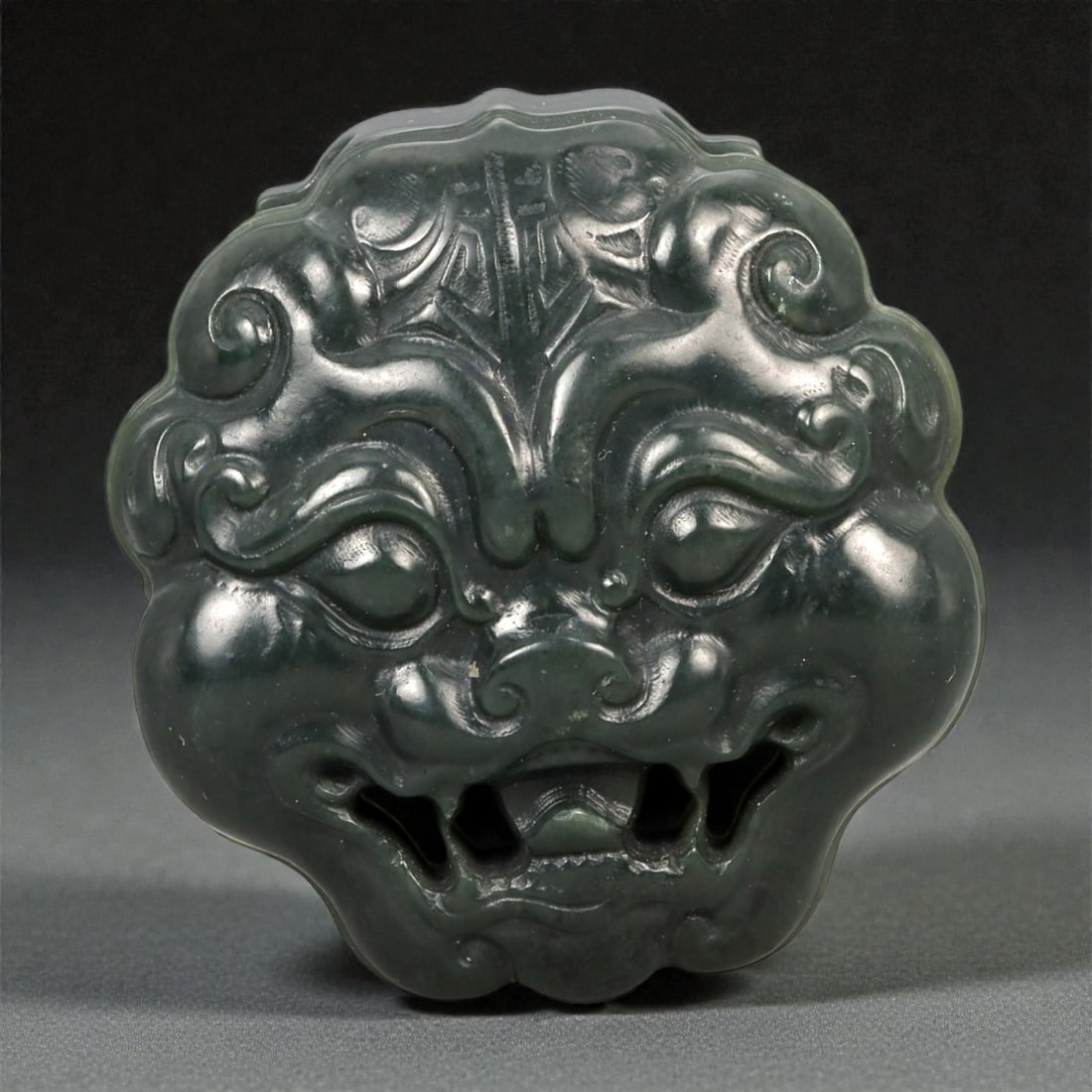 A Chinese Nephrite Jade Beast-Form Buckle, 20th Century, 55FB: Carved from dark green nephrite jade with subtle tonal variation and a lustrous polish, the buckle is modeled as a stylized mythical beast face rendered in high relief. The frontal composition is symm