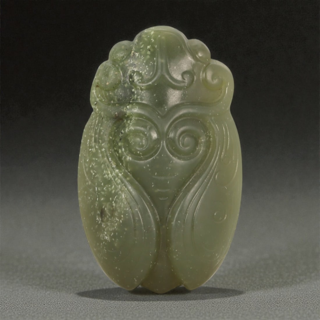 A Chinese Nephrite Jade Cicada Pendant, 20th Century, 99FB: Carved from olive-green nephrite jade with scattered light inclusions and areas of soft translucency, the pendant is worked in relief to depict a cicada in a compact, frontal composition. The head is