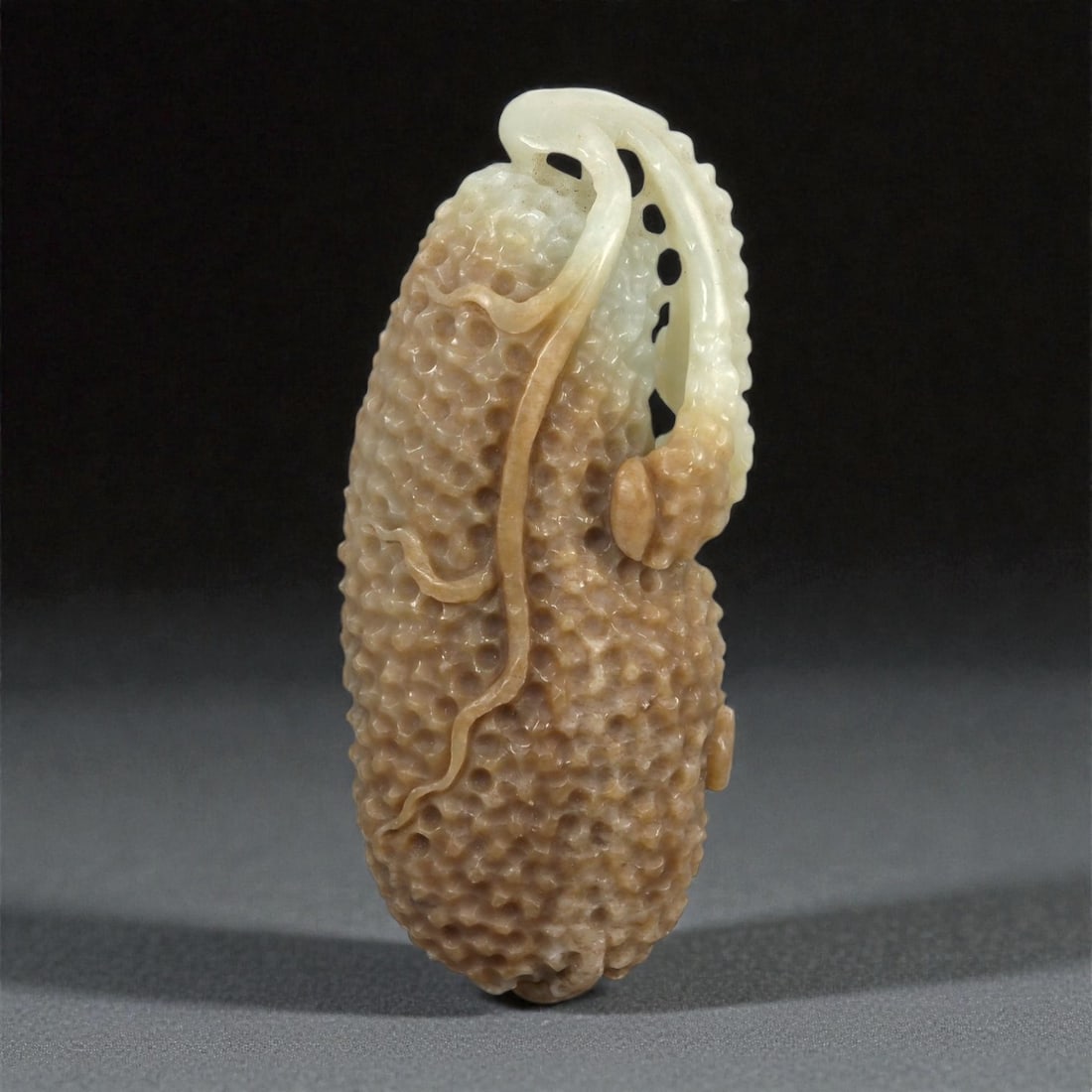 A Chinese Nephrite Jade Fruit-Form Pendant, 20th Century, 72FA: Carved from pale celadon to light brown nephrite jade with natural tonal variation and subtle translucency, the object is modeled in the form of an elongated fruit. The surface is densely worked with