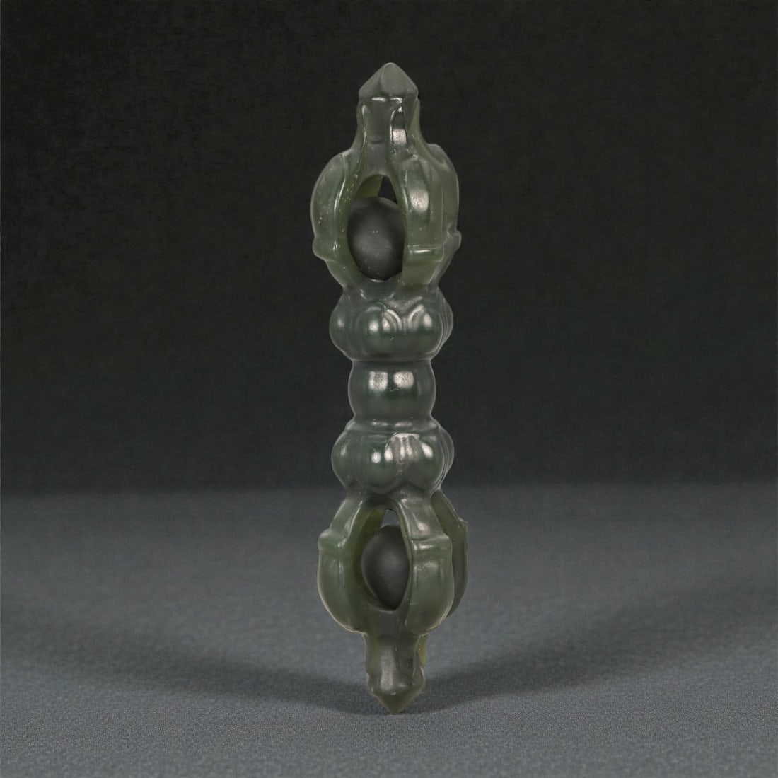 A Chinese Nephrite Jade Vajra (Dorje) Ornament, 20th Century, 8YA: Carved from dark green nephrite jade with areas of lighter translucency and a smoothly polished surface, the object is formed as a vajra (dorje) with symmetrical terminals aligned along a central axis