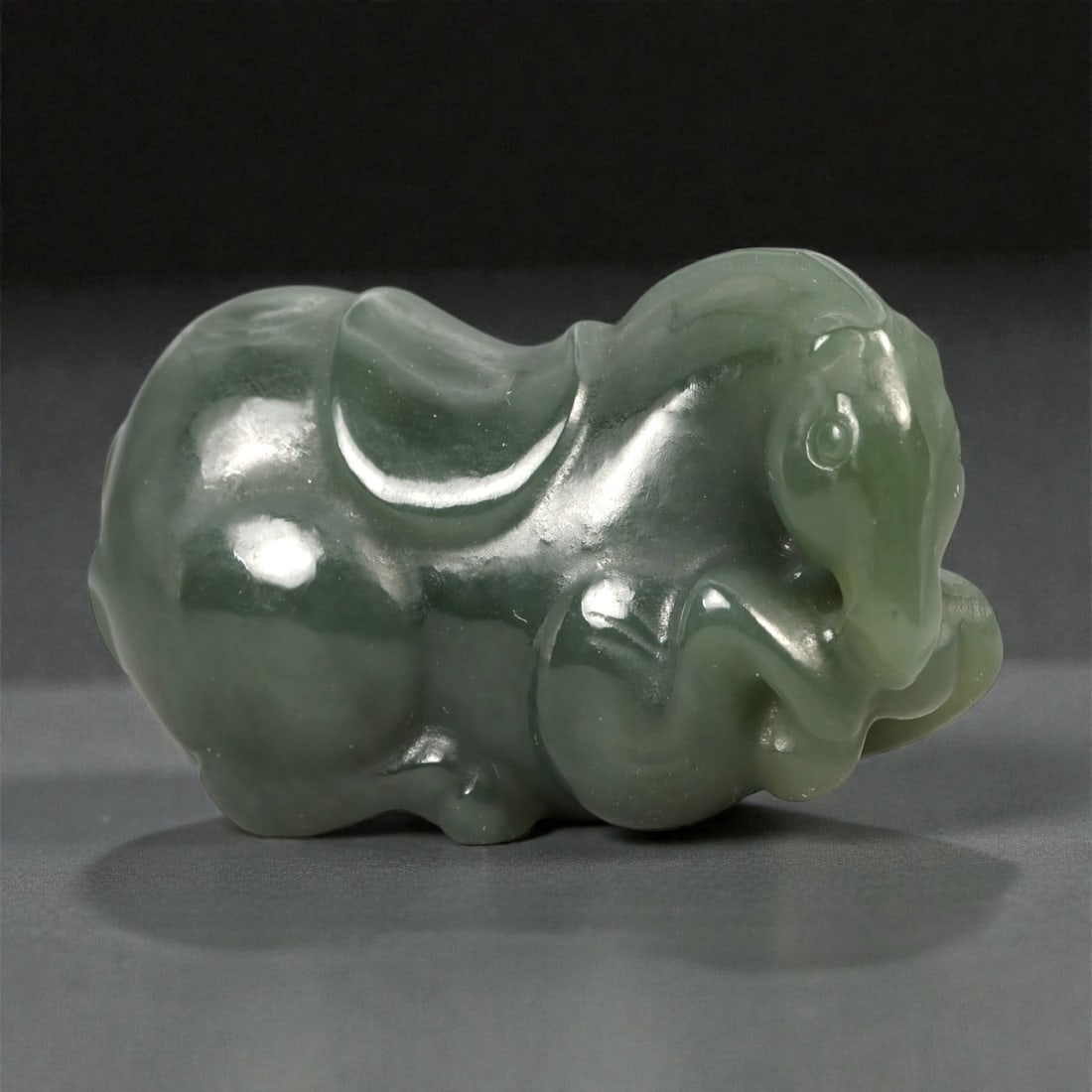 A Chinese Nephrite Jade Horse Figure, 20th Century, 93FA: Carved from dark green nephrite jade with areas of soft translucency and minor natural inclusions, the figure represents a horse shown recumbent with the legs folded beneath the body. The head is slig
