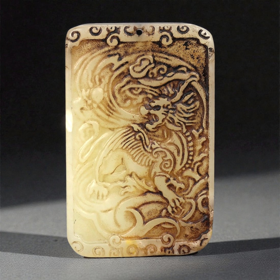 A Chinese Jade Dragon Plaque, 20th Century, 25FA: Carved from pale cream-colored jade with areas of brown surface staining and darker inclusions, the plaque is rectangular in form with rounded corners and worked in low relief on one face. The surface
