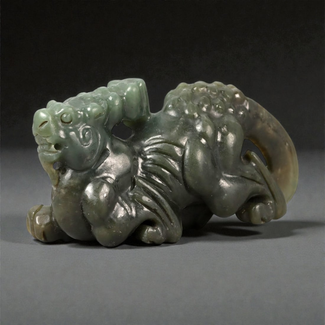 A Chinese Nephrite Jade Lion Figure, 20th Century, 85FA: Carved from dark green nephrite jade with areas of translucency and natural tonal variation, the figure represents a recumbent lion shown resting on all fours. The head is turned slightly to one side