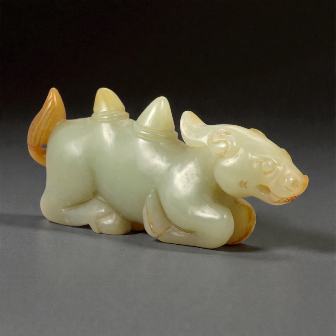 A Chinese Nephrite Jade Recumbent Mythical Beast, Qing Dynasty, 21FB: Carved from pale celadon nephrite jade with areas of warm russet skin retained along the extremities, the figure represents a recumbent quadruped modeled with a compact body and legs folded beneath. T