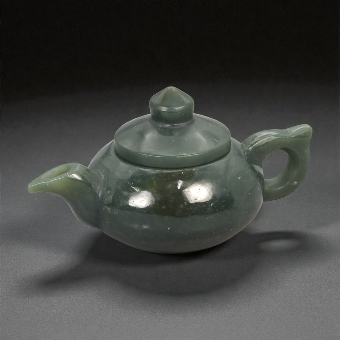 A Chinese Jade Teapot, 20th Century, 28YA: Carved from dark green jade with scattered lighter inclusions and natural translucency, the vessel is formed as a small teapot with a compressed globular body resting on a flat base. One side is fitte