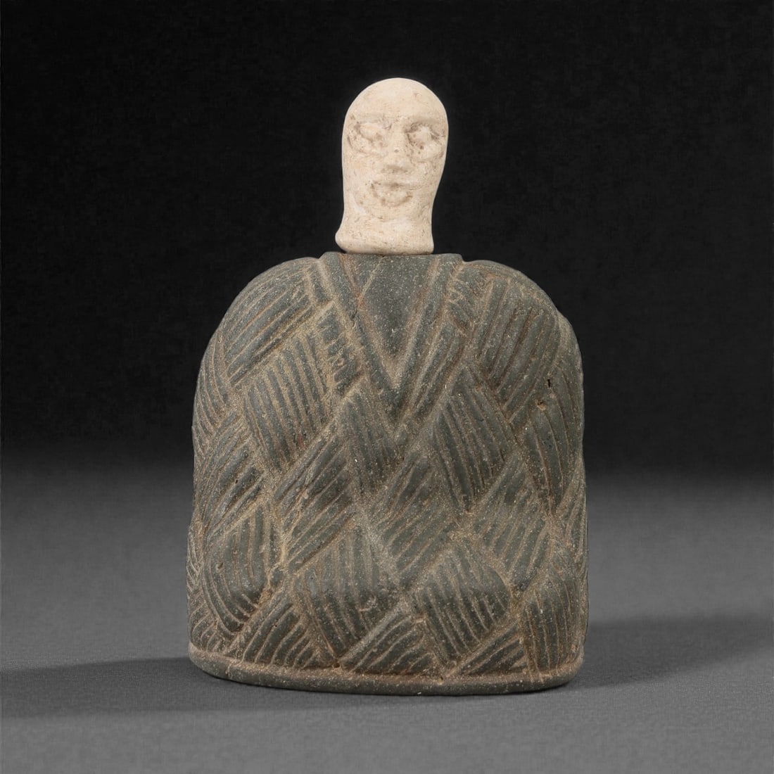 A Bactrian Composite Stone Idol, 3rd Millennium BCE, 34FD: Carved from dark gray stone forming a bell-shaped body, the surface is worked with deeply incised geometric patterns arranged in overlapping diagonal bands, creating a textured, cloak-like appearance.