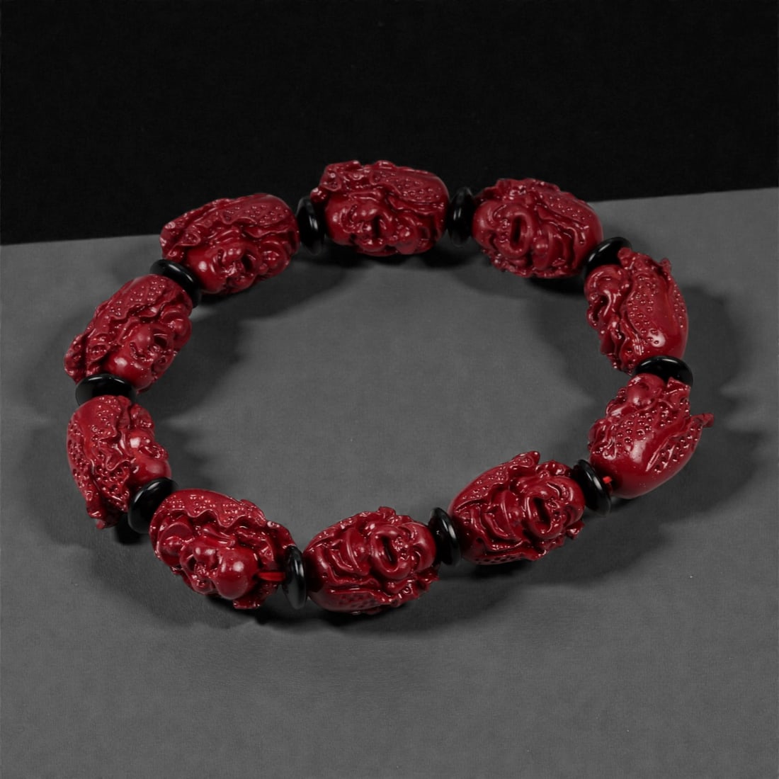 A Chinese Cinnabar Carved Bead Bracelet, 20th Century, 65FD: Composed of oval beads carved from cinnabar, a mercury sulfide material traditionally used in Chinese ornamental and ritual contexts. Each bead is centrally pierced and carved in high relief with dens