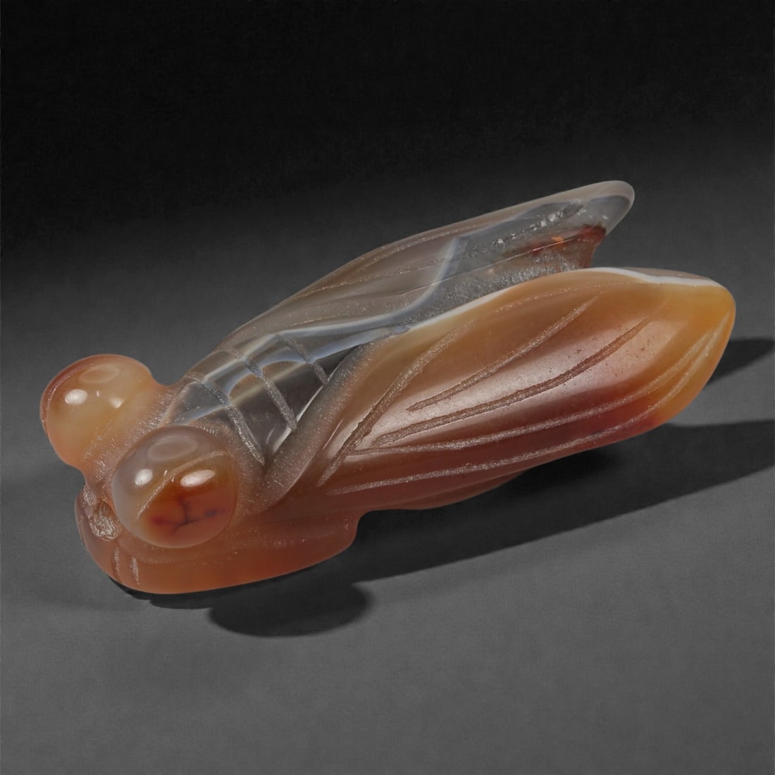 A Chinese Onyx Cicada Pendant, 20th Century, 46FD: Carved from banded onyx with natural tonal gradation ranging from pale grey and translucent white to warm brown and amber hues. The pendant is modeled in the form of a cicada, rendered in the round wi