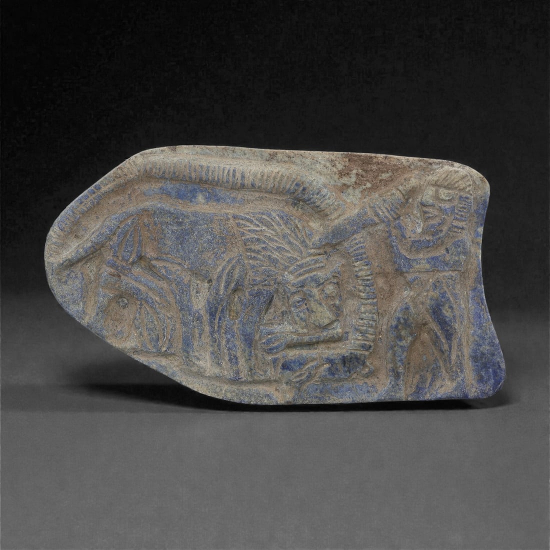 A Syro-Hittite Lapis Lazuli Plaque, Iron Age II, 68ZB: Carved from a mottled blue lapis lazuli slab, the plaque is worked in low relief with a figural scene arranged within a lightly raised border. At left appears a recumbent quadruped with prominent eye,