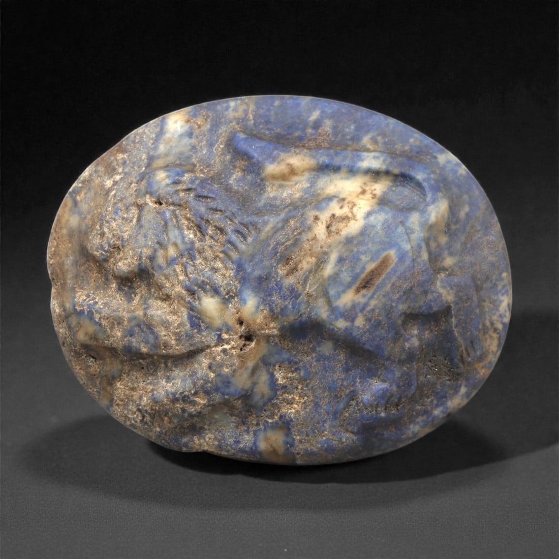 A Near Eastern Lapis Lazuli Lion Plaque, 3rd-2nd Millennium BCE, 16ZA: Carved from deep blue lapis lazuli with natural white and gold veining, the object is shaped as an oval plaque with a low-relief depiction of a recumbent lion. The head is shown in profile with a shor