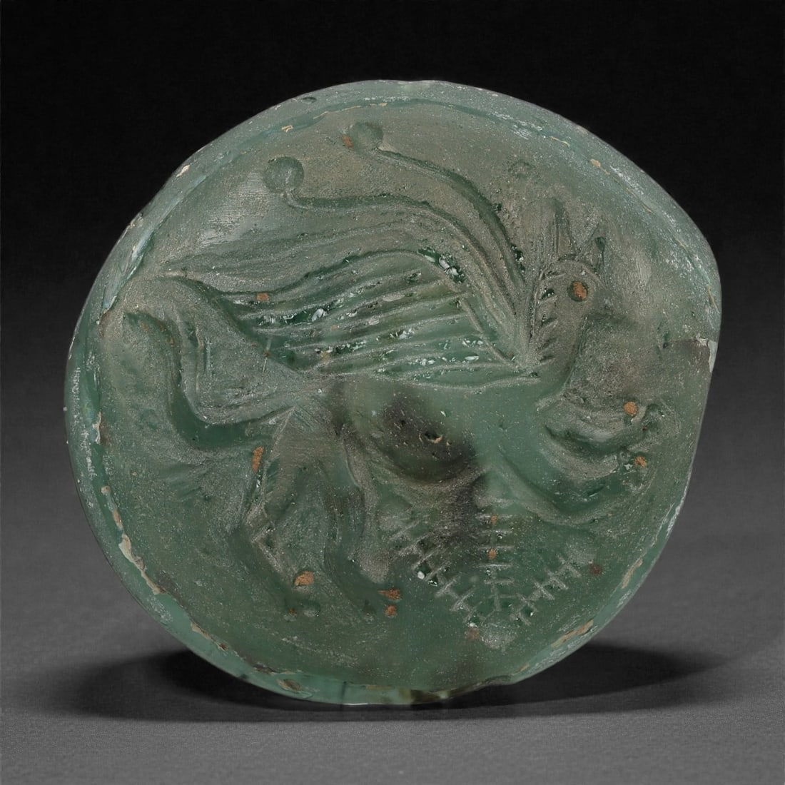 A Sasanian Green Glass Medallion with Winged Hybrid Creature, 3rd-7th Century CE, 28LP: Cast from dark green translucent glass, the medallion is circular in form and decorated in low relief with a winged hybrid creature shown in profile. The body is delineated by shallow grooves defining