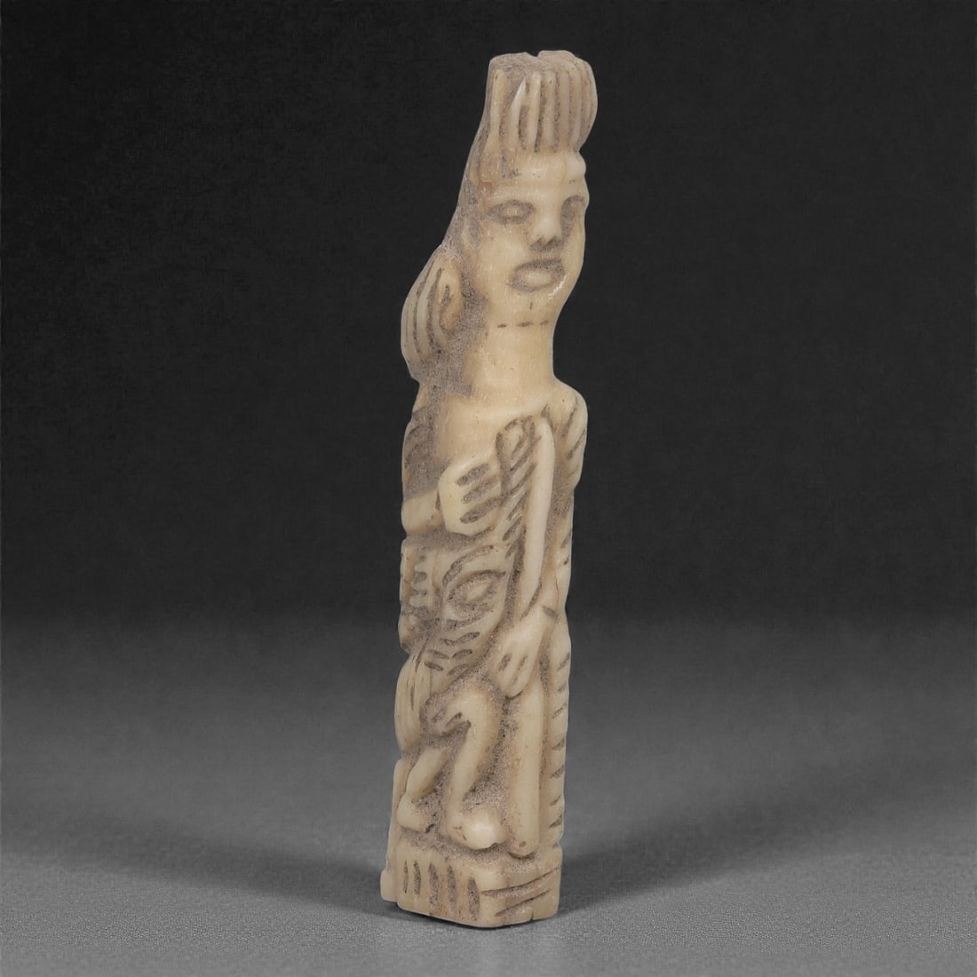 A Parthian Carved Bone Figurine, 1st-3rd Century CE, 3LJ: Carved from a light cream-colored bone with darker surface infill along the recessed areas, the figure is rendered in an elongated upright stance with a rectangular base. The head is marked by a tall,
