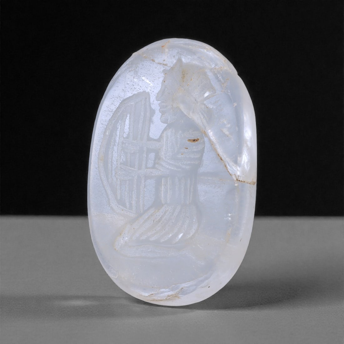 An Ancient Sasanian Glass Intaglio, 3rd-7th Century CE, 65L: Carved from translucent pale blue glass, the oval intaglio bears an incised representation of a standing or kneeling figure in profile, shown holding a rectangular object interpreted as a lyre or fram