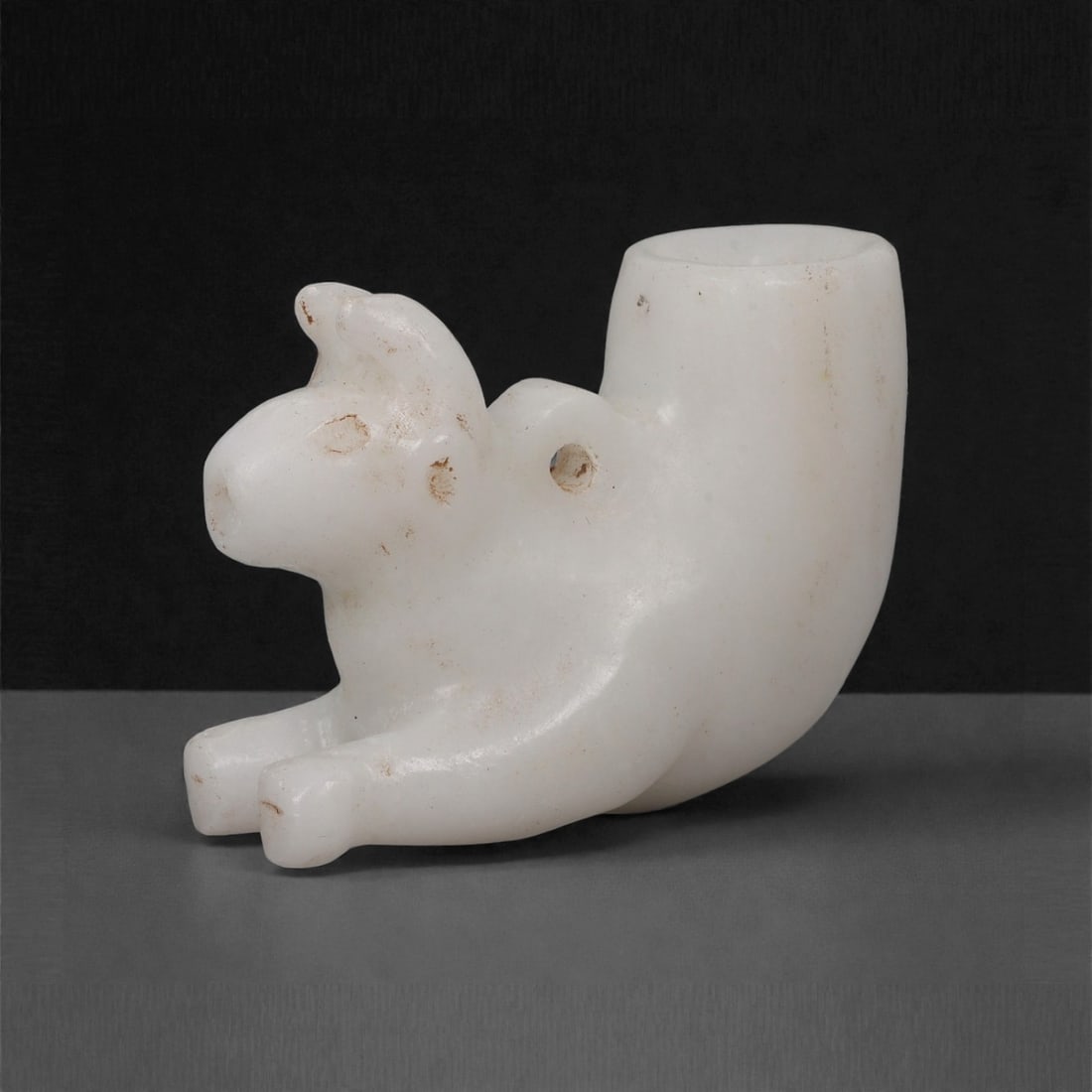 A Parthian Marble Zoomorphic Rhyton Terminal, 1st Century BCE-2nd Century CE, 42LP: Carved from fine white marble, the object is shaped as a recumbent quadruped with a rounded snout, small upright ears, and shallowly incised circular eyes. A perforation is drilled laterally through t