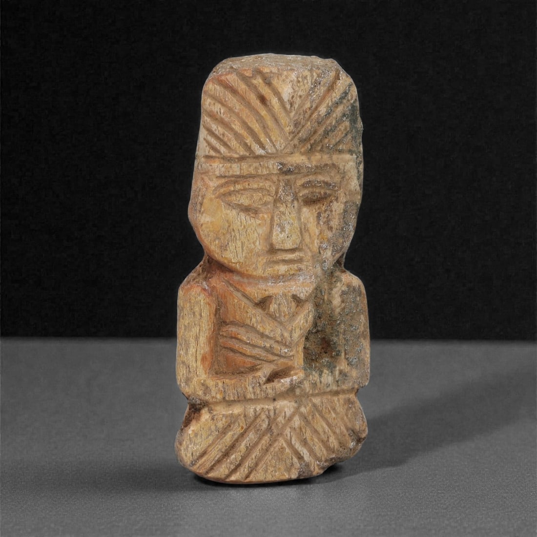 A Coptic Carved Bone Seated Figure, 5th-7th Century CE, 9LJ: Carved from a light brown bone with darker surface accretions, the figure is shown seated with the legs folded beneath and the hands positioned at the chest. The head is marked by a tall, striated hea
