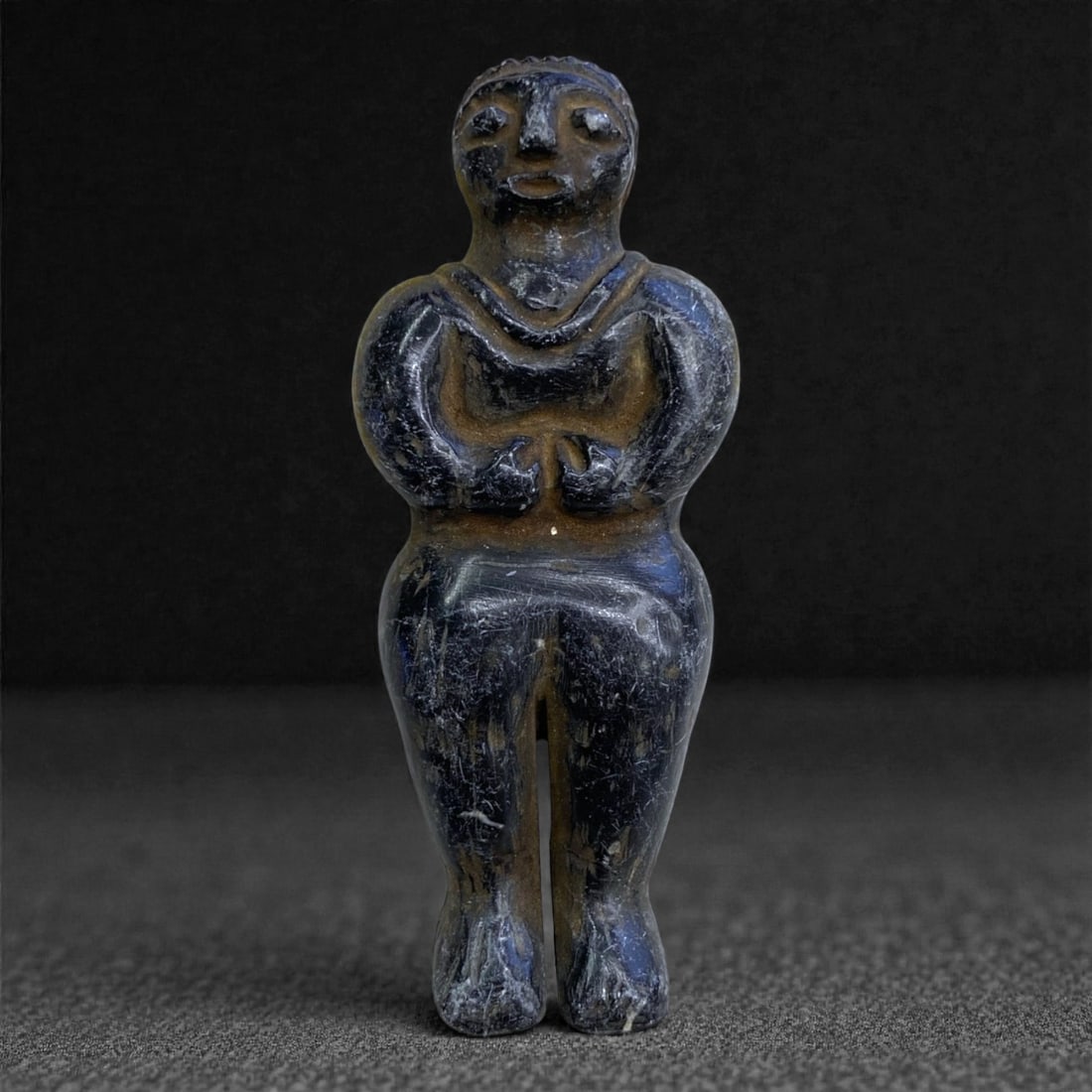 A Near Eastern Stone Anthropomorphic Idol, 2nd-1st Millennium BCE, 81C: Carved from a dark, fine-grained stone with areas of lighter surface abrasion, the figure is depicted standing with legs together and arms bent forward at the elbows, the hands meeting at the waist. T