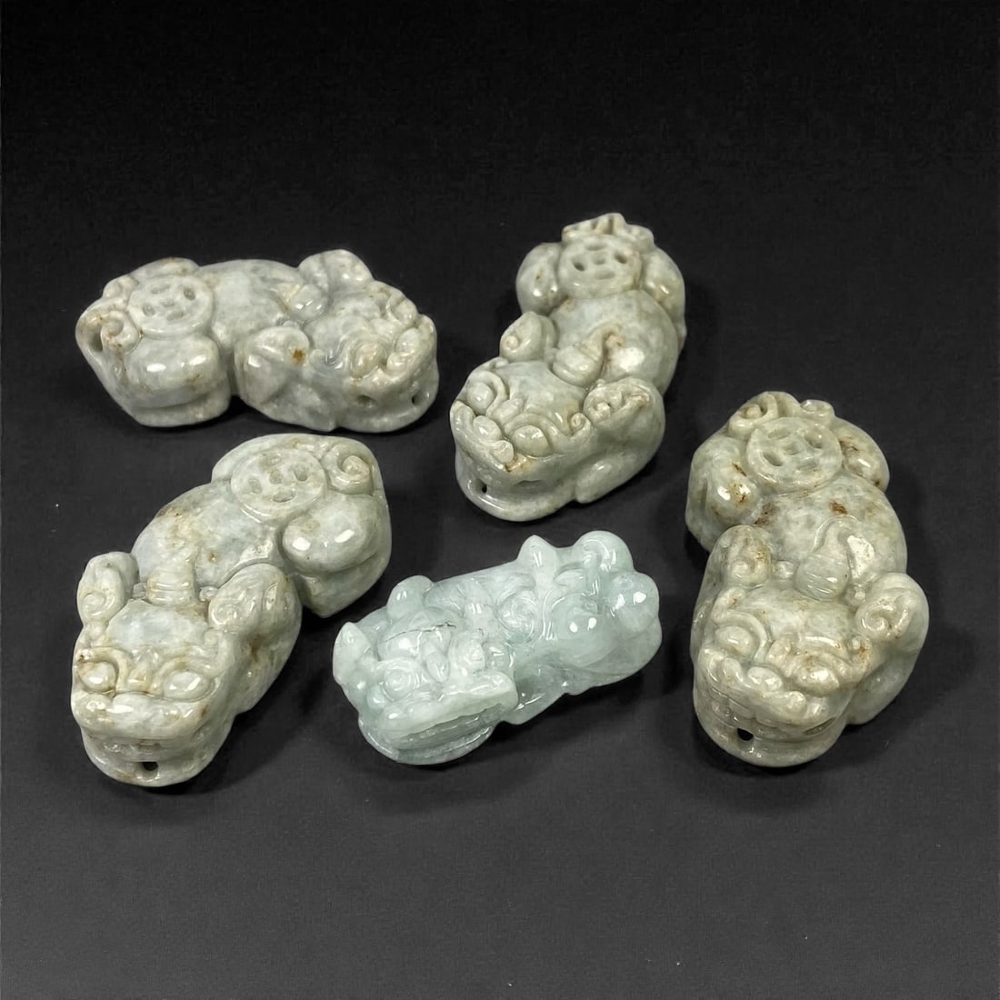A Group of Five Chinese Jadeite Jade Pixiu Figures, 20th Century, 5XJ: Comprising five small carved pixiu figures executed in pale green to grey-green jadeite jade with varying degrees of translucency and natural mottling, each modeled in a compact recumbent posture. The