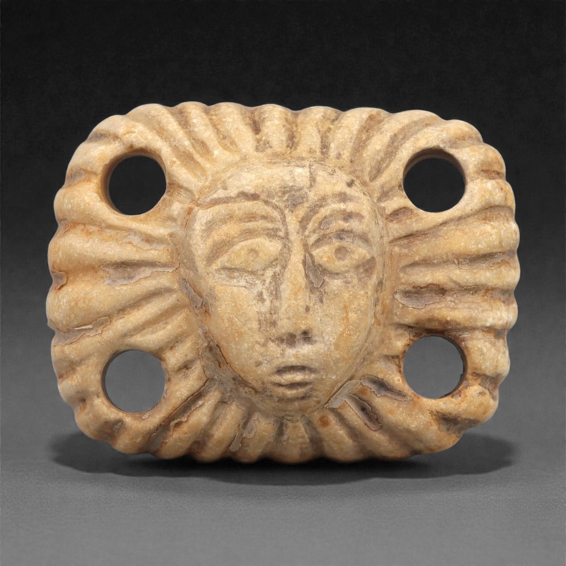 A Syro-Hittite Limestone Face Plaque, Early 1st Millennium BCE, 57LP: Carved from light beige limestone with natural surface veining, the plaque presents a frontal human face enclosed within a radiating border of fluted segments. The facial features are delineated with 