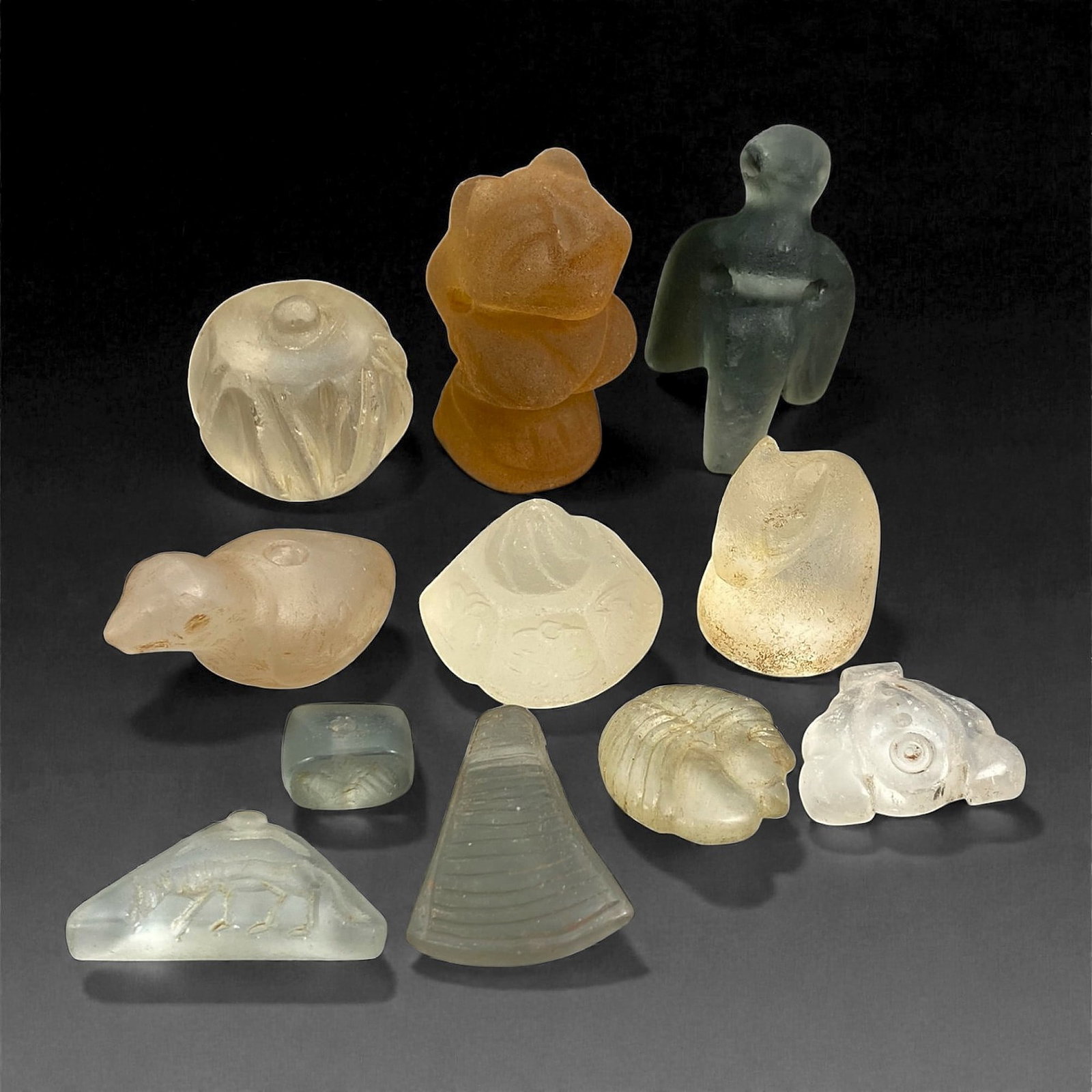 A Group of Medieval Glass Amulets and Relics, Circa 10th-14th Century, 80V: Comprising eleven small glass objects formed from translucent to semi-opaque glass in pale green, clear, amber, and honey tones, with visible weathering, surface pitting, and iridescence consistent wi