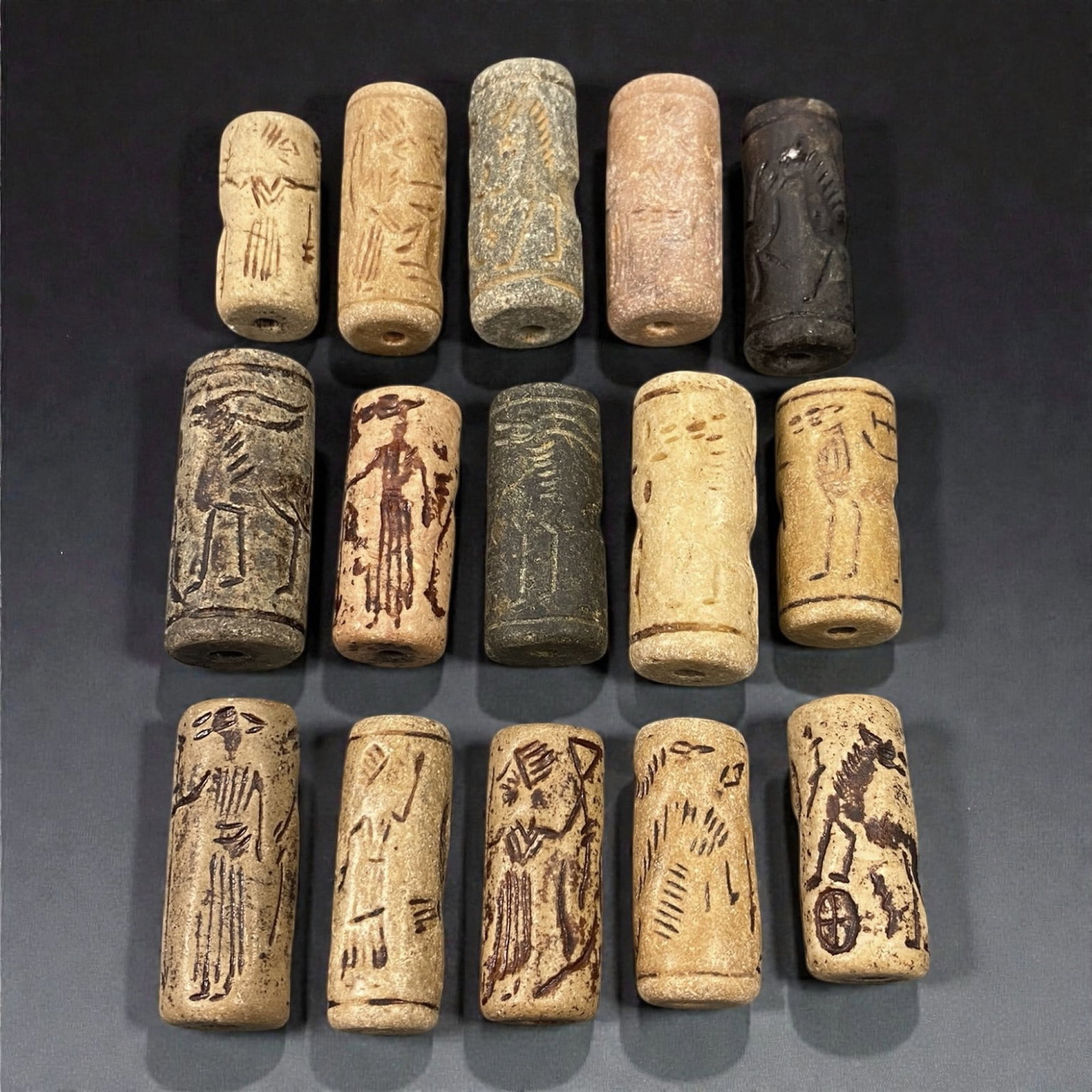 A Group of Near Eastern Stone Cylinder Seals, Mesopotamia, 3rd-2nd Millennium BCE, 39W: Comprising fifteen cylindrical seals carved from a range of stone materials, including buff, beige, grey, and dark stone with natural inclusions and surface variation. Several examples are engraved wi