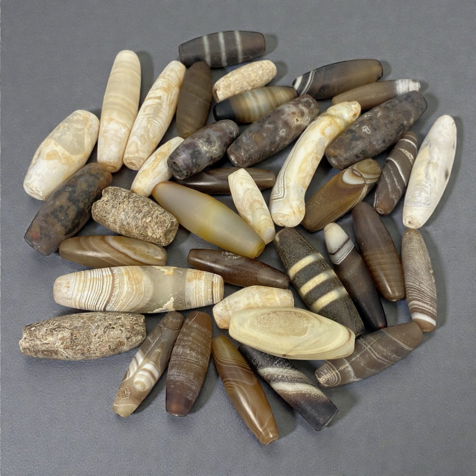 A Group of Ancient-Medieval Agate Beads, Circa 1st Millennium BCE, 13W: The group comprises thirty-seven agate and chalcedony beads of elongated tubular, barrel, biconical, and oval forms, carved from banded and translucent stone in tones of cream, beige, brown, grey, bla