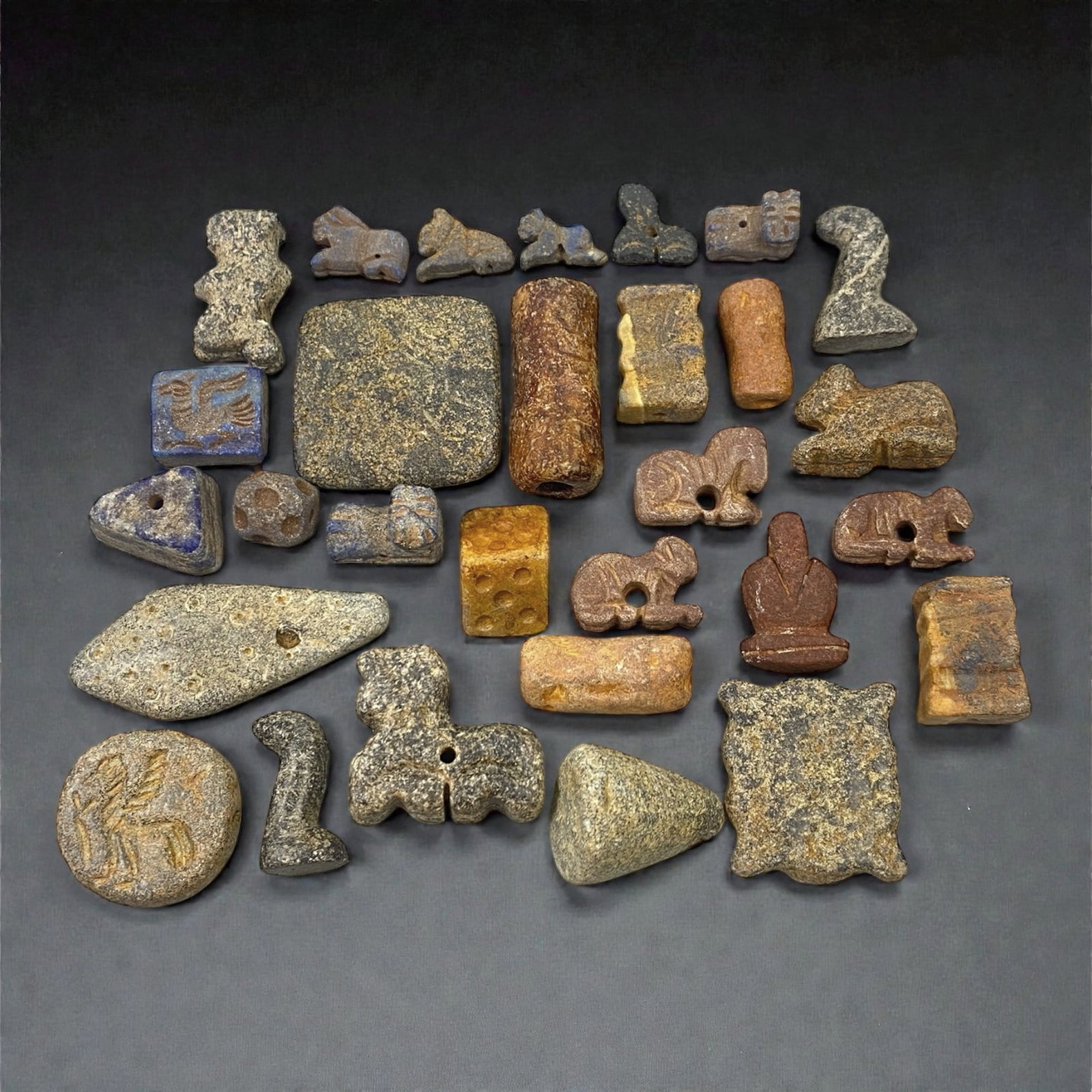 A Group of Bactrian Stone Beads and Amulets, 3rd-2nd Millennium BCE, 31W: Comprising a heterogeneous group of beads and small amuletic elements carved from stone of varied composition and coloration, the assemblage includes geometric, zoomorphic, and stylized anthropomorphi